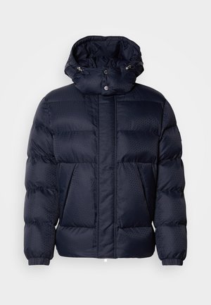 Tumedast puffer jakk, millel on tekstuuriga viimistlus, kõrge krae, eemaldatav kapuuts ja kaks esikäsi. Omab esist veni ja nööpe.