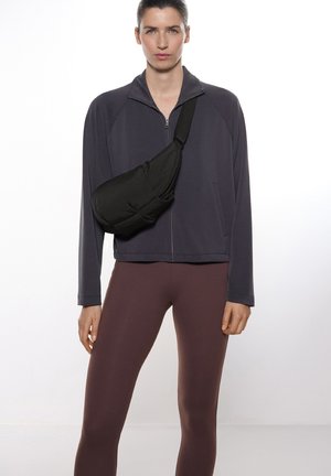 Schwarze Umhängetasche diagonal über einer grauen Zip-Jacke getragen, kombiniert mit dunkelbraunen Leggings. Glattes Material, minimales Design, funktioneller Stil.