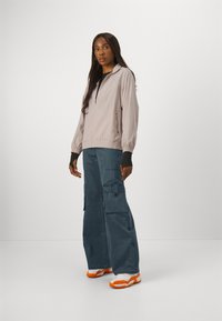 Veste zippée rose clair avec des poignets élastiques, associée à un pantalon cargo bleu foncé avec des poches latérales et une coupeLoose. Les chaussures sont blanches avec des accents orange.