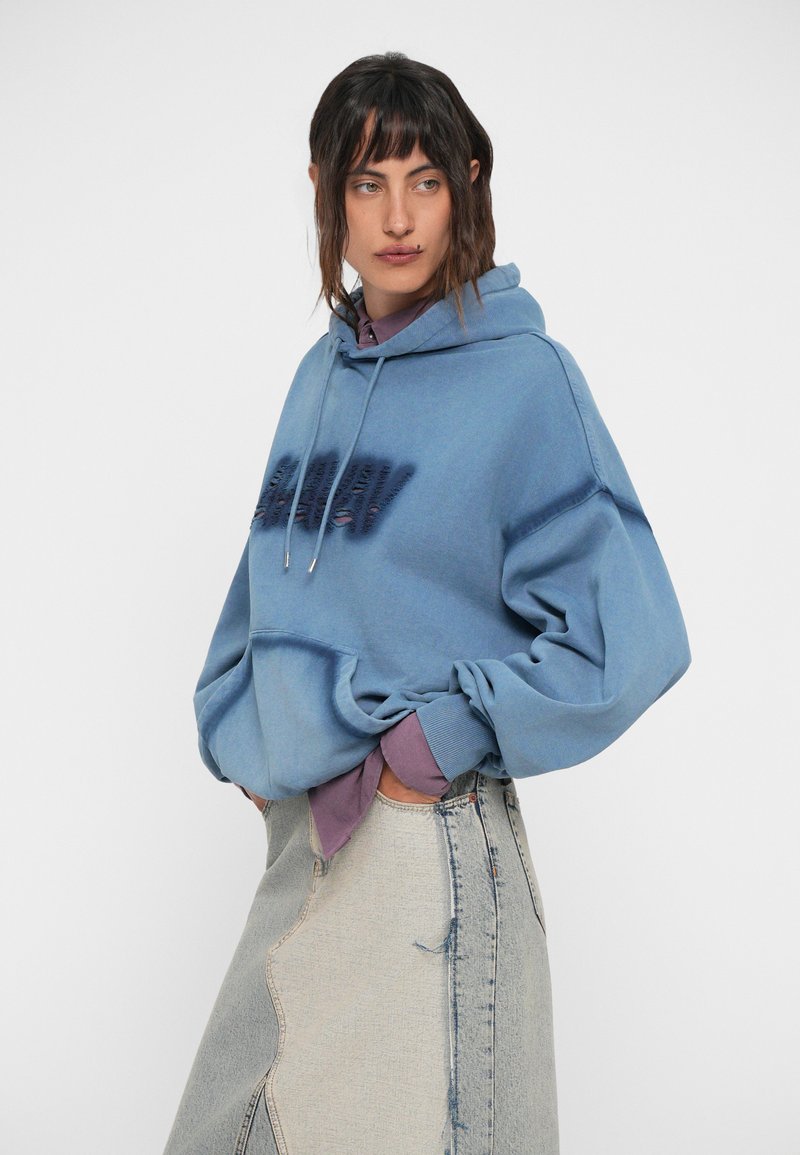 Lyseblå oversized hoodie med en lomme på forsiden, snorhue og mørk tekstdesign. Bær over en lilla skjorte og et denimskørt.