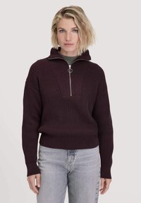 Burgunderfarbener Strickpullover mit hohem Kragen, einem Reißverschluss vorne und geripptem Textur. Kombiniert mit hellgrauen Jeans.