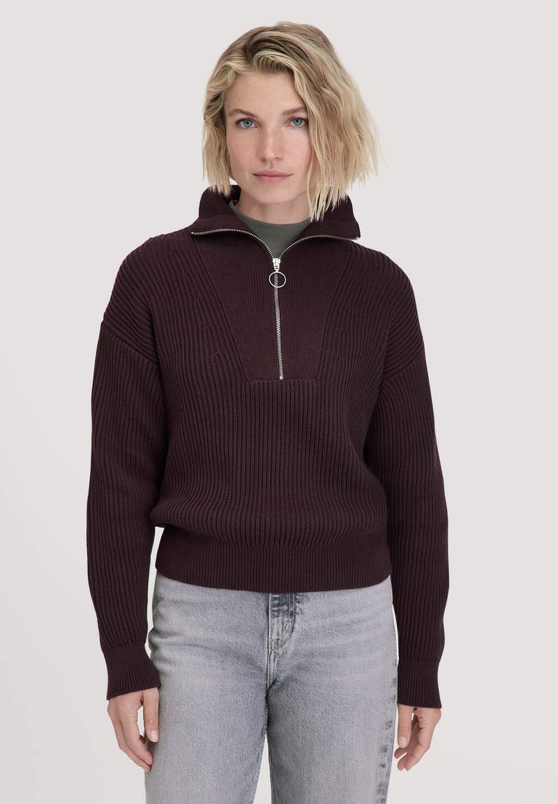 Burgunderfarbener Strickpullover mit hohem Kragen, einem Reißverschluss vorne und geripptem Textur. Kombiniert mit hellgrauen Jeans.