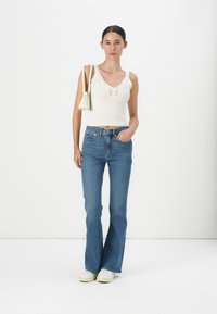 Hvit strikket croptopp med v-hals og mønstret tekstur, kombinert med blå vidde jeans. Modellen bærer en liten hvit håndveske.