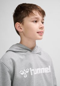 Grauer Hoodie aus weichem Stoff, mit einem weißen Logo auf der Brust. Das Design umfasst eine vordere Tasche und eine Kapuze mit Kordel.