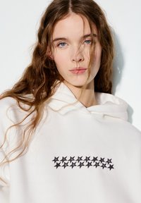 Weißer Hoodie mit einem schwarzen Sternenmuster auf der Vorderseite, das in einer Linie angeordnet ist. Weicher Stoff mit lockerem Sitz und Kapuze mit Kordelzug.
