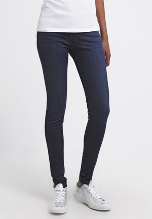 Jeans oscuros, ajustados, de mezclilla, con una textura suave y un diseño simple, combinados con zapatillas plateadas.