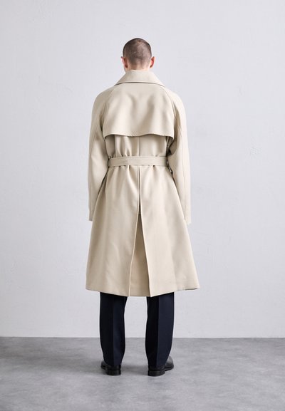 Persona con i capelli corti che indossa un trench coat beige, pantaloni neri e scarpe nere, in piedi di fronte a un muro grigio chiaro semplice.