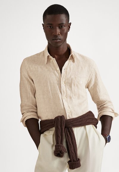 Νεαρός άνδρας που φοράει μπεζ ριγέ πουκάμισο με κουμπιά, με τα μανίκια σηκωμένα, εκρού παντελόνι, καφέ πουλόβερ δεμένο στη μέση και ρολόι χειρός στο αριστερό καρπό.