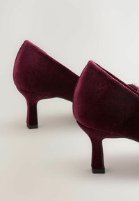 Par de zapatos de tacón medio de terciopelo burdeos con tacones cuadrados, exhibidos sobre una superficie clara.