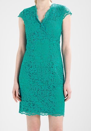 Femme portant une robe en dentelle turquoise à manches courtes, avec un col en V festonné et un motif floral, debout devant un fond uni.