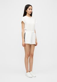 Λευκό κοντομάνικο μπλουζάκι με στρογγυλή λαιμόκοψη, συνδυασμένο με λευκά σορτς με κορδόνι στη μέση και μαύρες ρίγες στα πλαϊνά. Φορεμένο με λευκά αθλητικά παπούτσια.