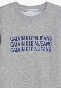 Szary sweatshirt z prążkowanym dekoltem i powtarzającym się napisem "CALVIN KLEIN JEANS" w wyraźnym granatowym kolorze. Miękki materiał tekstylny o melanżowej fakturze.