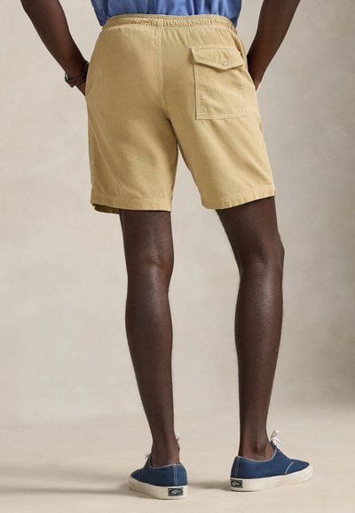 Άτομο που φοράει μπεζ σορτς βελουτέ με πίσω τσέπη και μπλε πάνινα παπούτσια, στέκεται μπροστά σε ουδέτερο φόντο.