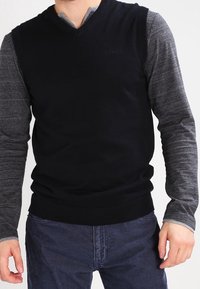 Svart v-ringad tröja med ribbad nederkant, överlagd på en grå långärmad skjorta. Tyget verkar mjukt och visar en lagerdesign med minimal varumärkesprofil.