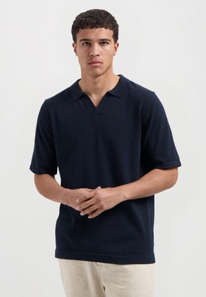 Marineblauwe polo met korte mouwen en een V-hals, met opgerolde mouwen. Gebreide textuur en een ontspannen pasvorm; eenvoudig ontwerp zonder patronen of accenten.