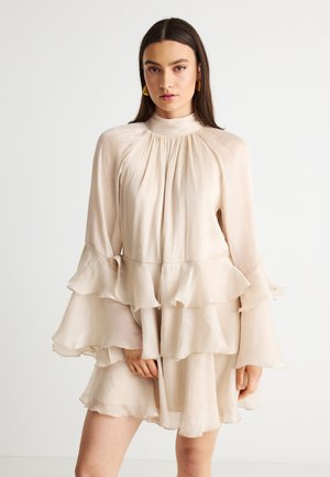 Femeie purtând o rochie bej din chiffon stratificat, cu mâneci lungi evazate și cercei aurii cu formă geometrică, pe un fundal alb simplu.