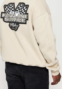Sweatshirt beige avec logo graphique "Fastlane Service" et drapeaux à damier au dos. Comprend des poignets et un ourlet côtelés. Porté avec un pantalon noir.