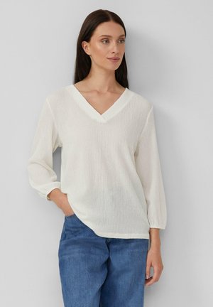 Vrouw in een witte, gestructureerde V-hals langemouwtop met elastieken manchet en blauwe spijkerbroek, staand voor een effen lichtgrijze achtergrond.