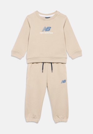 Beige leggingsættet til småbørn med sweatshirt med lange ærmer og elastiske manchetter, der har blå New Balance-logoer på brystet og bukserne.