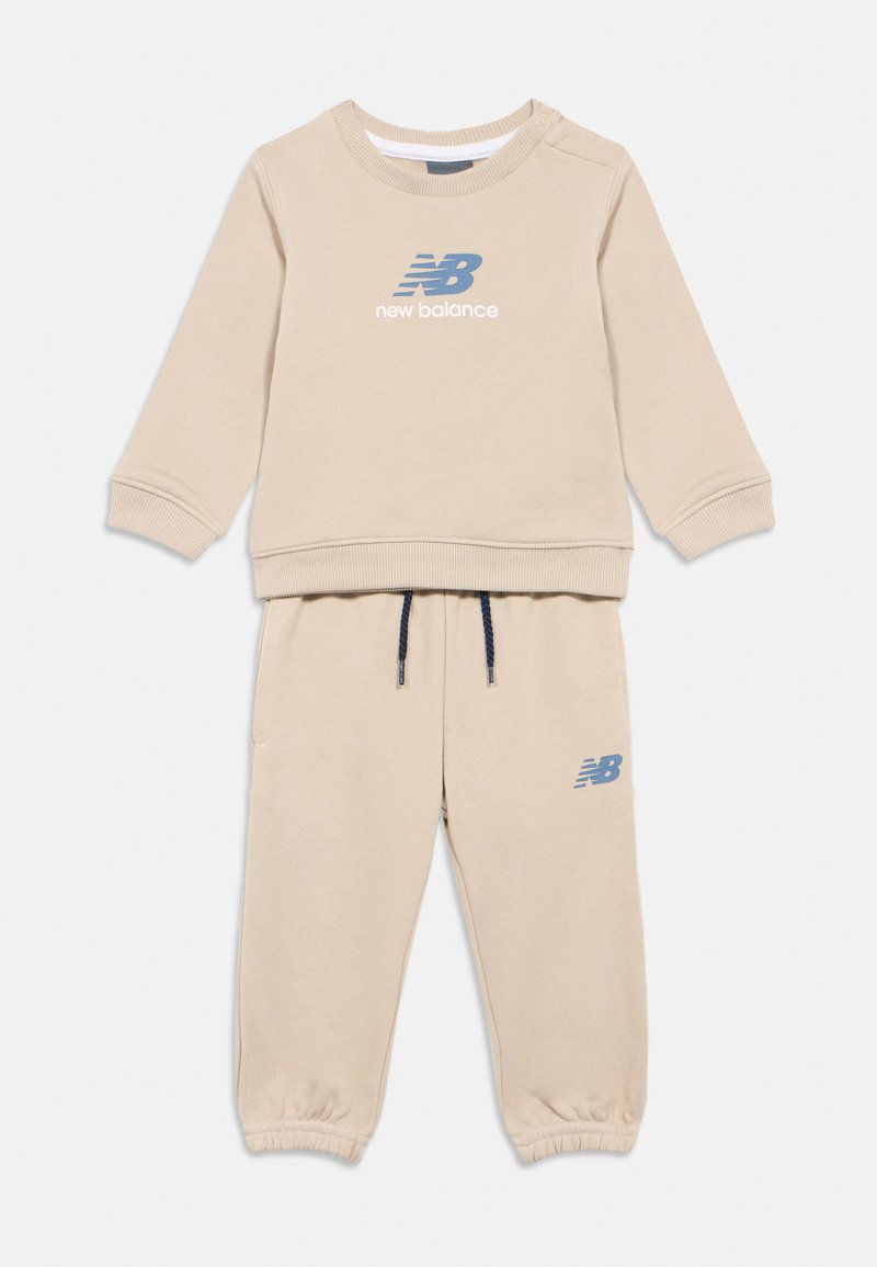 Tuta beige per bambini con felpa a maniche lunghe e polsini elasticizzati, con loghi blu di New Balance sul petto e sui pantaloni.