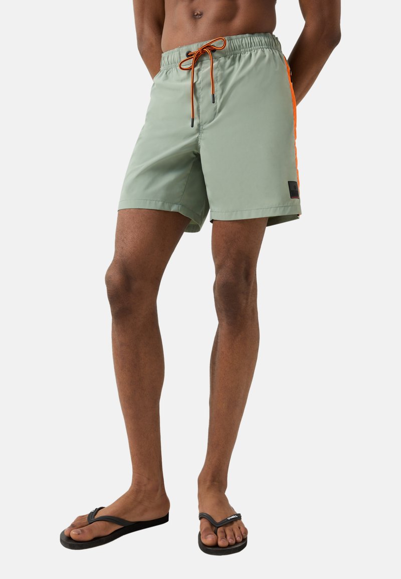 Homme portant un short de bain vert clair avec des bandes latérales orange et un cordon de serrage orange, assorti de tongs noires, debout sur un fond blanc.