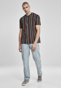 Fekete rövid ujjú póló függőleges piros, sárga és zöld csíkokkal. Világoskék farmernadrággal és szürke tornacipővel párosítva.