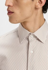 Kahviin värjätty vaaleankerman värinen nappipaita, jossa on punaisia ja ruskeita geometrisia kuvioita. Ominaisuuksiin kuuluu klassinen kaulus ja valkea nappisuljenta.