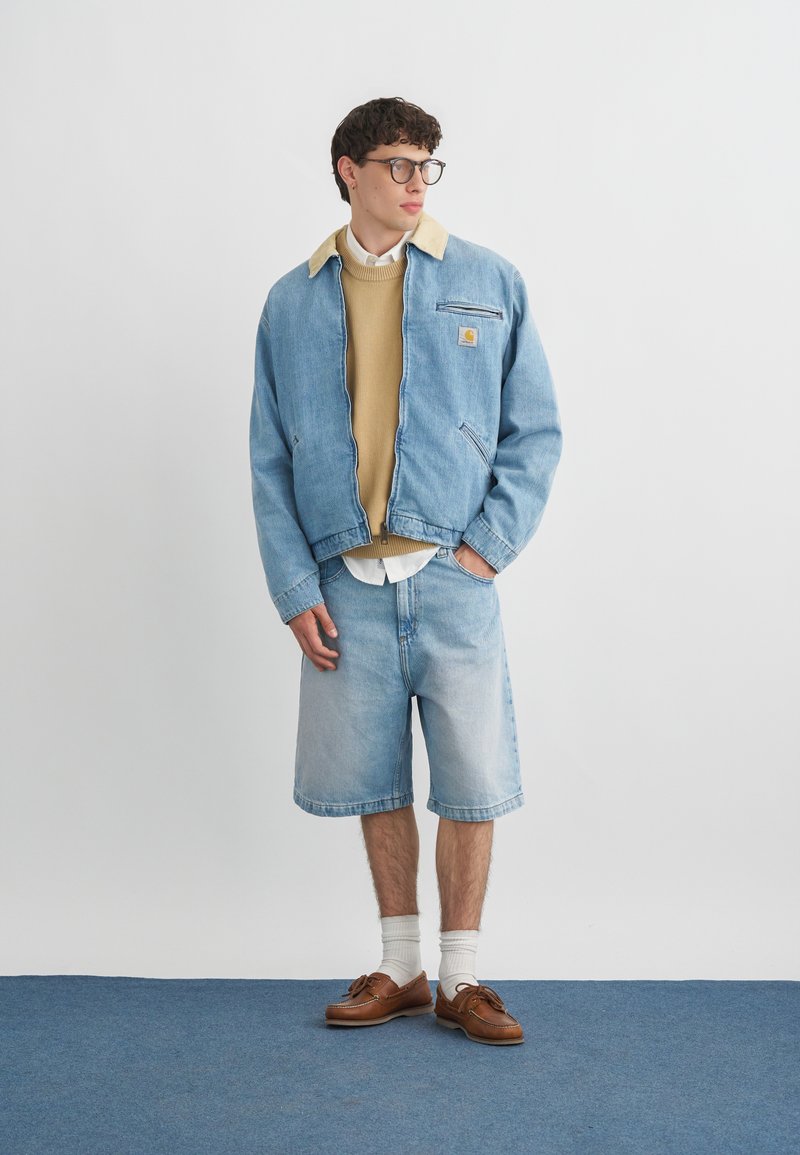 Jonge man met bril, beige trui onder een lichtblauw spijkerjasje en korte broek, bruine bootschoenen met witte sokken, staand op een blauwe vloer.