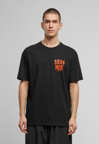T-shirt en coton noir à manches courtes, avec un texte graphique orange vif "SICK MODE" sur le côté gauche. Coupe décontractée et col ras du cou.