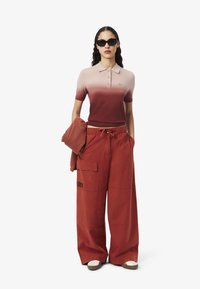 Polo rose avec un dégradé de rose clair à rouge profond, assorti à un pantalon rouge à jambes larges, doté de poches et d'un cordon de serrage. Lunettes de soleil noires.