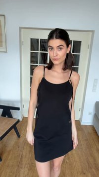 Femme aux cheveux foncés attachés, portant une robe noire simple sans manches, debout à l’intérieur sur un sol en bois près d’une porte vitrée.