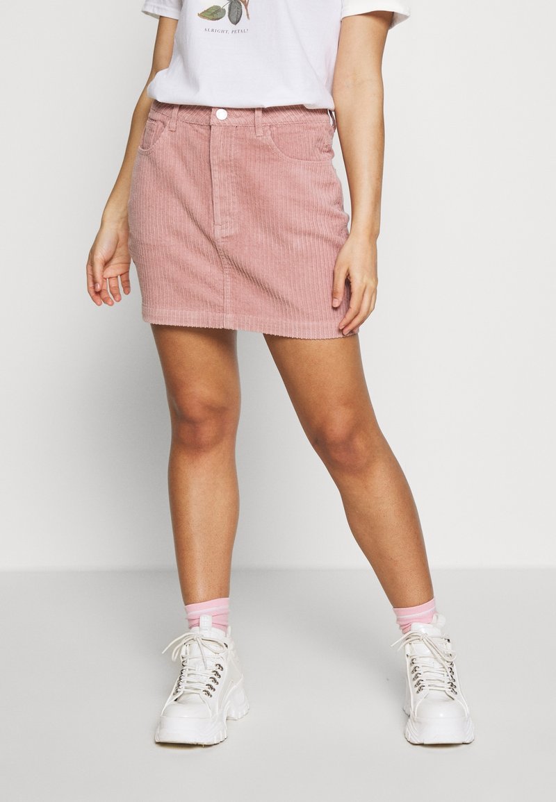 Femme portant une mini-jupe en velours côtelé rose, un T-shirt blanc à motif, des chaussettes roses et des baskets blanches à plateformes épaisses, debout devant un fond blanc.
