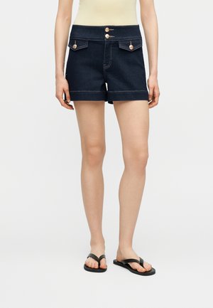 Nő sötét farmer rövidnadrágban, elülső zsebekkel és arany gombokkal, fekete papucsokkal és világos felsővel párosítva.