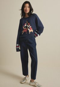 Femme portant un pull marine avec des chiens festifs en chapeaux de Noël et un jean sombre, se tenant avec une main dans sa poche contre un fond uni.