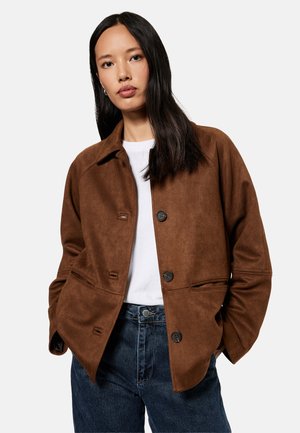 Frau mit langen dunklen Haaren, die eine braune, geknöpfte Jacke über einem weißen Hemd und blauen Jeans trägt, steht mit den Händen in den Taschen.