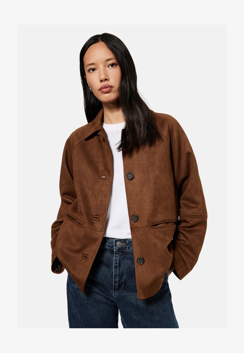 Woman with long dark hair wearing a brown buttoned jacket over a white shirt and blue jeans, standing with hands in pockets.