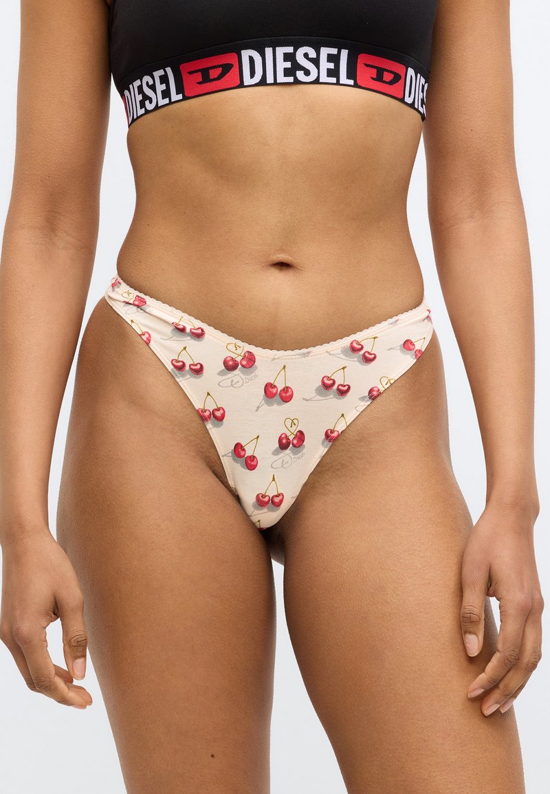 Beige thong with red cherry print, lace trim, and logo detail. Designed for a fitted silhouette with minimal coverage. Cotton blend fabric.