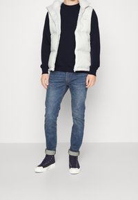 Persona che indossa un gilet bianco sopra un maglione nero, jeans blu arrotolati e sneakers alte navy, in piedi davanti a uno sfondo semplice.