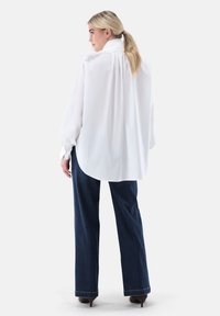 White long-sleeve blouse with a high collar and pleated back, paired with dark blue flared jeans and brown heeled shoes.