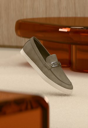 Mocassino grigio in camoscio con suola bianca e dettaglio fibbia argento, mostrato sospeso sopra uno sfondo di tappeto chiaro e mobili in legno.