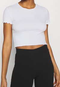 Biała krótka koszulka z okrągłym dekoltem i falbaniastymi krótkimi rękawami. Wykonana z gładkiego materiału, z asymetrycznym wykończeniem dołu.