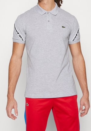 Hombre con polo de manga corta gris claro con cinta negra y blanca con logo en las mangas y pantalones cortos deportivos rojos con un pequeño logo verde.