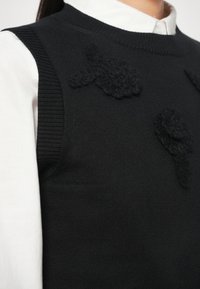 Pull en tricot noir avec des accents floraux texturés, associé à une chemise blanche à col en dessous. Dispose d'un col rond et d'ourlets côtes.