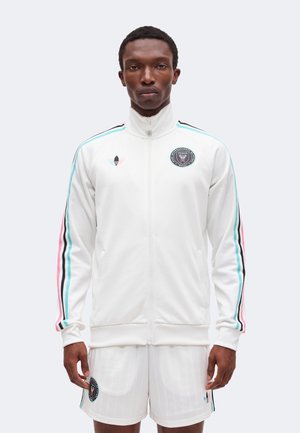 Mann trägt weiße Jacke mit Reißverschluss und passende Shorts mit Logos des Miami-Fußballteams und pink-blau-schwarz gestreiften Ärmeln.