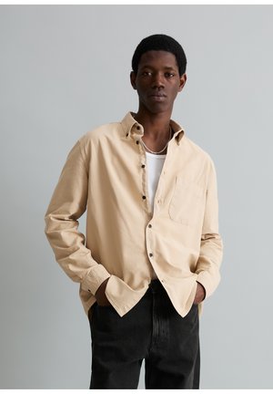 Junger Mann mit den Händen in den Taschen, trägt ein beiges Hemd über einem weißen Tanktop und schwarzen Hosen vor einem schlichten grauen Hintergrund.