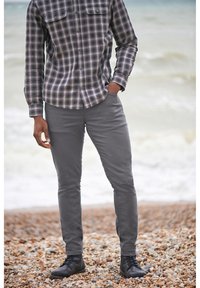 Plaid shirt with gray and black tones, two chest pockets, paired with gray tapered pants and black lace-up boots on a pebbled beach.