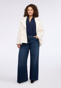 Cappotto doppiopetto color crema, top blu navy e jeans a gamba larga blu scuro. Scarpe nere appuntite completano il look. Tessuto liscio, taglio moderno.