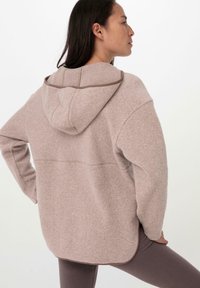 Pullover con cappuccio in morbido pile beige con dettagli delle cuciture in contrasto marroni, caratterizzato da una vestibilità comoda e un orlo posteriore leggermente più lungo.