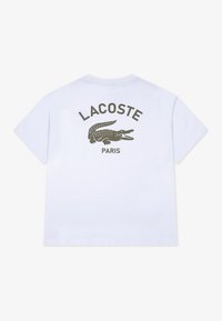 Biela tričko s logom krokodíla Lacoste v tmavozelenej farbe, s nápisom "LACOSTE" a "PARIS" vytlačeným nad logom na chrbte.
