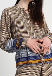 Bluse mit Knopfverschluss, einem geometrischen Muster in Braun und Gold, mit blauen Akzenten und kragenfreiem Ausschnitt. Lange Ärmel mit taillierten Manschetten.
